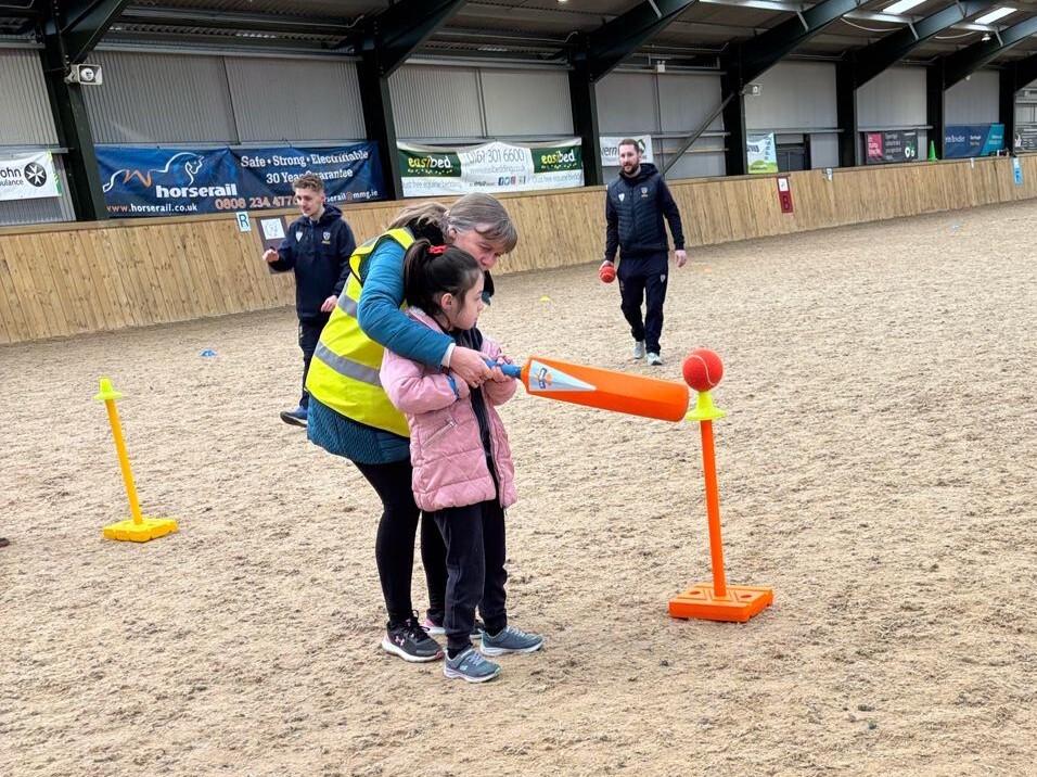 A young person learning how to hit a cricket ball with a cricket bat inside an indoor horse menage used as a venue for our Inclusion School Games events. The young person is being supported by an adult and two cricket instructors are seen in the background.